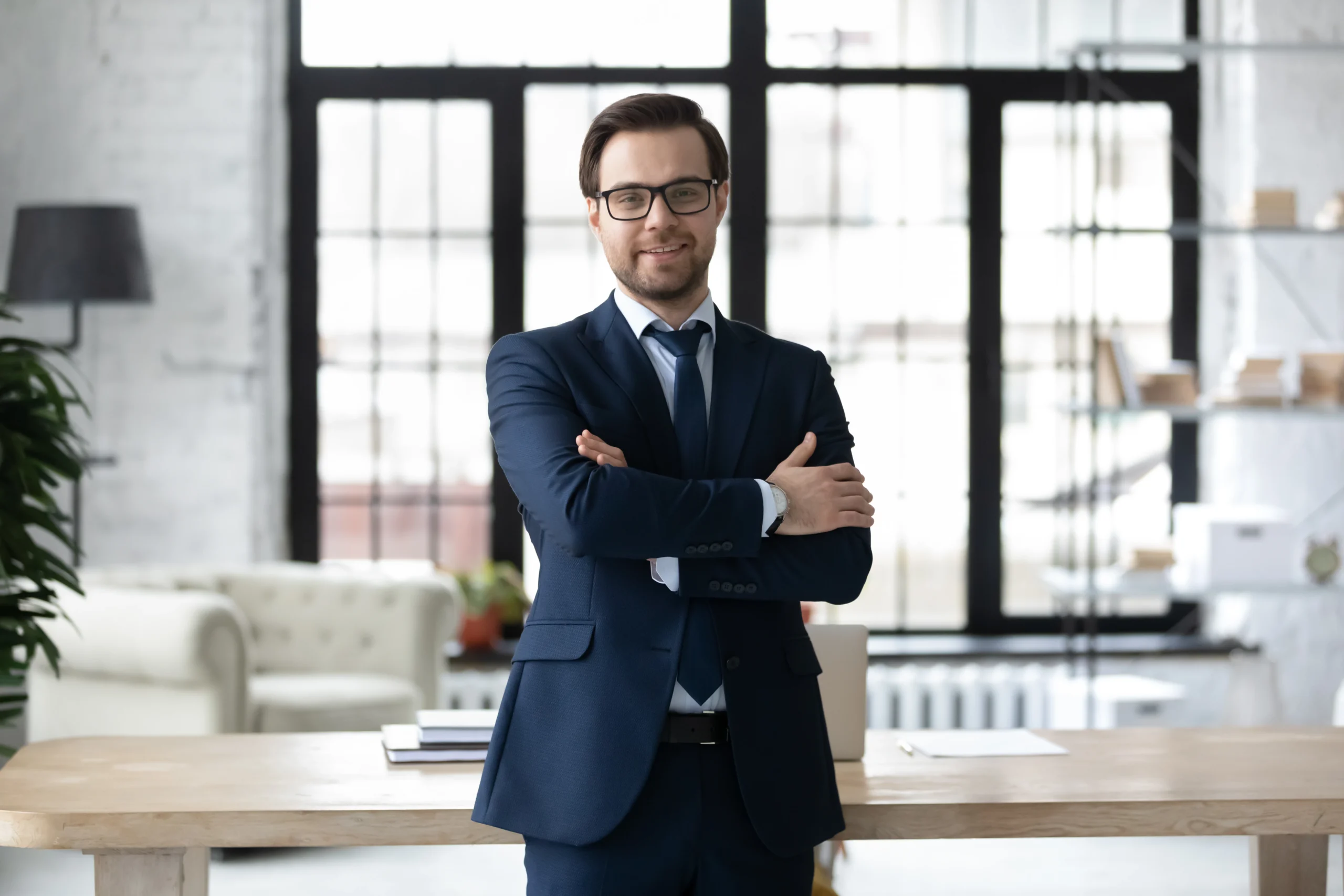 Portrait of smiling successful Caucasian businessman in formal suit glasses stand posing in modern office, happy young male boss or CEO look at camera, show confidence and power, leadership concept