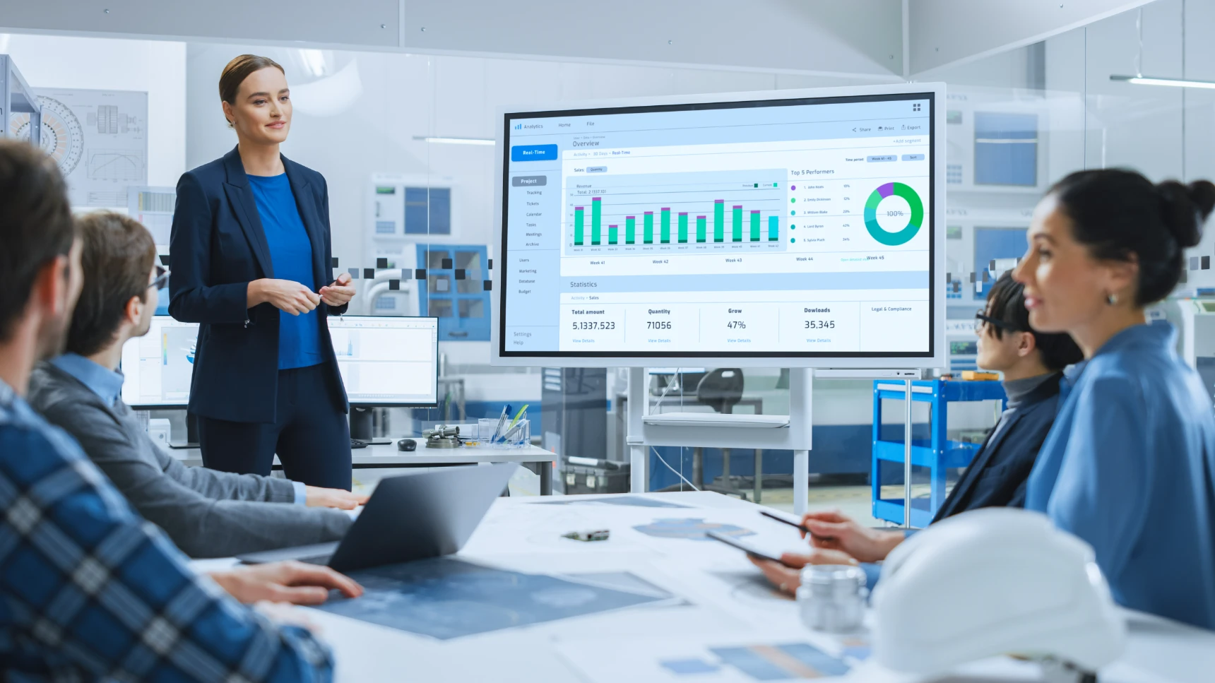 Modern Industrial Factory Meeting: Confident Female Engineer Uses Interactive Whiteboard, Makes Report to a Group of Engineers, Managers Talks and Shows Statistics, Growth and Analysis Information